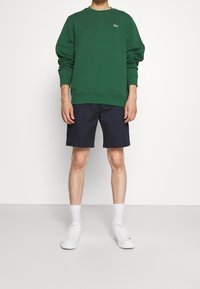 Zelená bavlněná mikina s žebrovanými manžetami a lemem, doplněná tmavě modrými šortkami. Model nosí bílé tenisky a kotníkové ponožky.