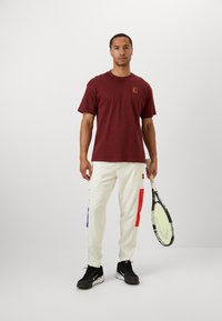 Maroon-burgundinine puuvillane t-särk, mille rinnal on väike logo, koos kreemika värviga pükstega, millel on punased ja sinised külgpanelid. Mustad tenniseketsid ja tennisereket.