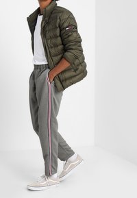 Puffjacka i olivgrön, vit t-shirt, grå sweatpants med röda och blå sidostreck, samt beige sneakers med vita sulor.