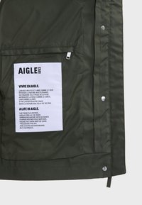 L'intérieur de la veste olive présente une étiquette blanche avec du texte. Le matériau semble lisse, avec une finition mate et des détails de couture subtils.
