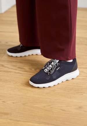 Zwarte mesh sport schoenen met witte zolen, voorzien van een patroon in elastische veters. Het ontwerp bevat een gladde donkere hakaccent.