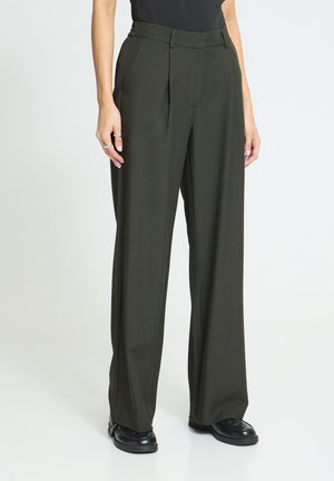 Personne portant un pantalon large couleur olive foncé, associé à des chaussures noires et un haut noir, debout sur un fond blanc.