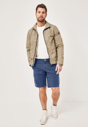 Bearded man wearing beige zip-up jacket, white t-shirt, navy cargo shorts, and white high-top sneakers, standing against a plain background.
