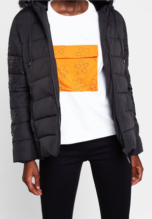 Osoba má na sobě černou prošívanou bundu přes bílou košili s velkou oranžovou kapsou zdobenou obrysovými květinovými vzory, doplněná černými kalhotami.