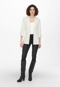 Blazer bianco con design sartoriale, apertura frontale e due tasche, abbinato a jeans skinny neri e stivali neri con tacco al ginocchio.
