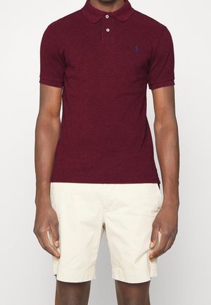Homme portant un polo bordeaux à manches courtes et un short beige clair arrivant aux genoux, debout devant un fond uni.