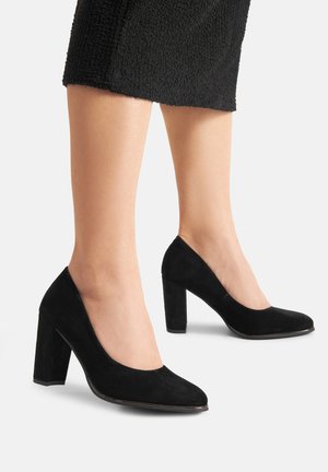 Schwarze Wildleder-Pumps mit hohem Absatz, abgerundetem Zehenbereich, klarer Form und quadratischem Absatz. Das Material hat eine weiche Textur.