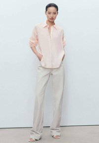 Camicia rosa chiaro a maniche lunghe con bottone frontale e dettagli in pizzo, abbinata a pantaloni larghi grigi chiari e sandali beige. Tessuto dalla consistenza liscia.