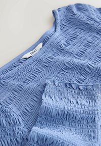 Haut bleu clair sans manches avec smocks, texture élastique froncée et une étiquette blanche portant la mention NEXT sur un fond clair.