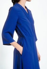 Plava obučena haljina od glatkog materijala s rukavima do tri četvrtine, V-izrezom i bočnim džepovima. Ima čistu, krojenu siluetu.