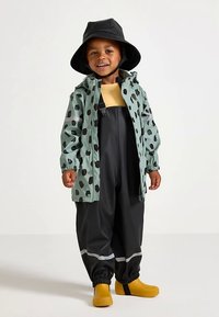 Vihreä sadetakki mustilla pilkuilla, keltainen paita, mustat vedenkestävät haalarihousut ja keltaiset sadetossut. Ominaisuuksiin kuuluu joustavat hihansuut ja heijastinnauhat.