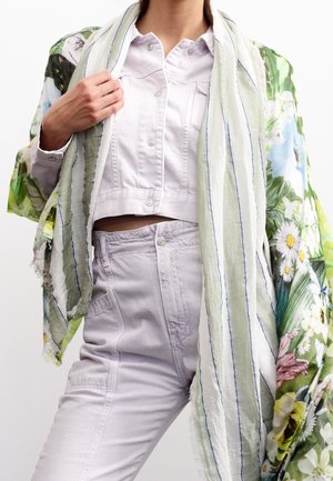 Femme portant une veste en denim violet clair et un pantalon assorti, avec une écharpe à motif floral drapée sur les épaules et une écharpe rayée devant.