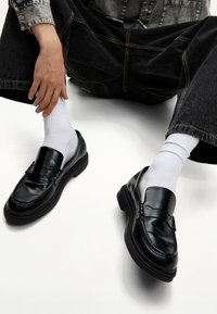 Black leather loafers with a smooth finish, featuring a raised seam detail, paired with white crew socks and worn with dark baggy jeans.