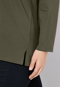 Chemise à manches longues vert olive avec une fente sur le côté et une texture douce, associée à un jean en denim foncé. Les détails de couture sont visibles.