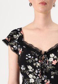 Vrouw in een zwarte bloemenjurk met kantafwerking en korte mouwen, waarbij haar schouders zichtbaar zijn en ze zilveren ringoorbellen draagt.