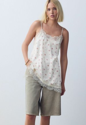 Junge Frau mit blondem Haar trägt eine weiße, mit Spitze verzierte, geblümte Camisole und beigefarbene knielange Shorts und posiert mit einer Hand in der Tasche.