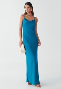 Turkizna maxi obleka iz satena z naramnicami iz tankih trakov in izrezom v obliki lasulje. Oblečena s teksturirano belo torbico in bež sandali.