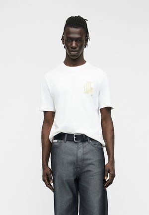 Νεαρός άνδρας με πλεξούδες, φορώντας ένα λευκό μπλουζάκι με τύπωμα, μπλουζάκι μέσα σε σκούρα γκρι τζιν με μαύρη ζώνη, στέκεται μπροστά σε απλό φόντο.