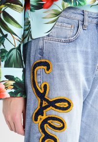 Top de color azul claro con estampado floral, combinado con jeans de mezclilla claros que presentan grandes letras bordadas en amarillo y marino con purpurina.
