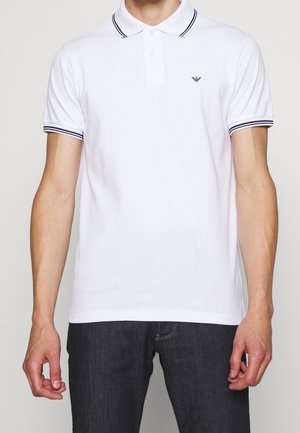 Homme portant un polo blanc à manches courtes avec un col et des manches rayés bleu marine, associé à un jean foncé, debout devant un fond uni.