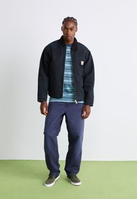 Black, cotton jacket with a collar and front zipper, layered over a blue and white striped shirt. Paired with dark pants and green sneakers.