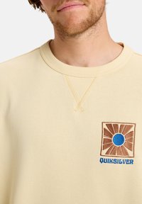 Light yellow sweatshirt with a round neck, featuring a brown and blue embroidered logo and square design on the left chest. Textured fabric.