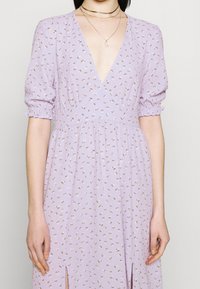 Robe florale lilas avec un décolleté en V plongeant, des manches courtes bouffantes et une taille froncée. Comprend des motifs floraux subtils et des fentes sur les côtés.