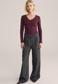 Burgunderfarbener langärmliger V-Ausschnitt-Pullover, kombiniert mit hoch geschnittenen, weit geschnittenen grauen Hosen mit einem gesprenkelten Muster und einer Knopfdetaillierung an der Vorderseite.