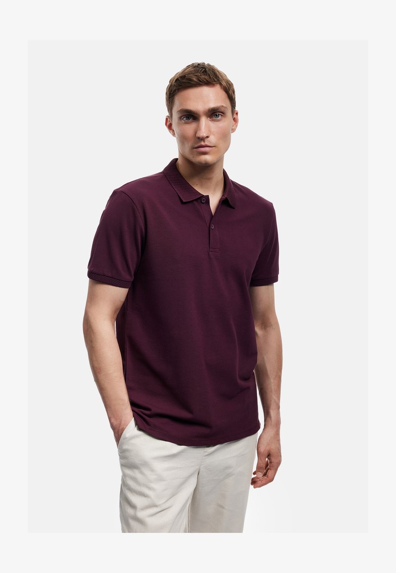 Junger Mann trägt ein kurzärmeliges, dunkelweinrotes Polohemd und helle beige Hosen, steht mit einer Hand in der Tasche vor einem schlichten weißen Hintergrund.