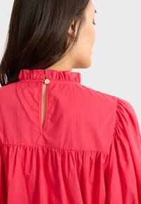 Blusa de algodón roja con un escote con volantes, detalle de abertura en la espalda y mangas fruncidas. Textura suave, tejido ligero y diseño casual.