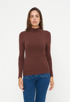 Femme portant un pull à col roulé brun côtelé ajusté et un jean bleu, debout sur un fond blanc uni.
