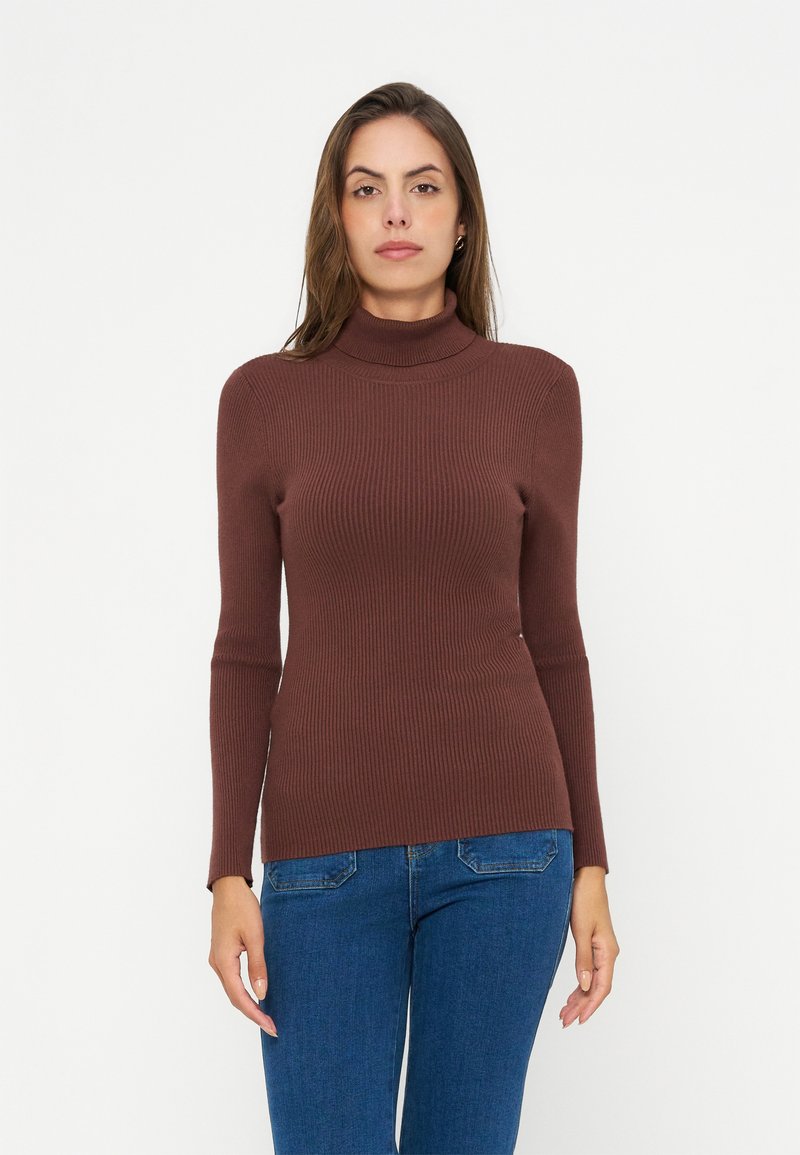 Femme portant un pull à col roulé brun côtelé ajusté et un jean bleu, debout sur un fond blanc uni.