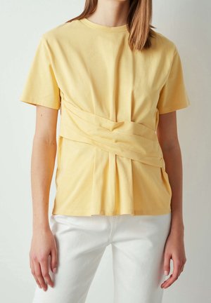 Kvinde iført en gul top med korte ærmer og et bredt omslagsdetalje ved taljen samt hvide bukser, stående foran en ensfarvet baggrund.