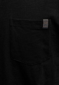 Μαύρη υφασμάτινη τσέπη σε ρούχο με μια μικρή γκρι και πορτοκαλί ετικέτα «Jack & Jones» ραμμένη στην πάνω άκρη της τσέπης.