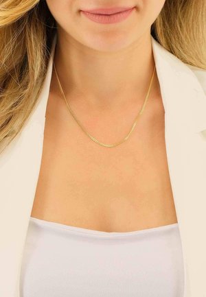Femme portant un collier en or fin sur un top blanc sans bretelles et un blazer beige, avec des cheveux blonds lâches visibles aux épaules.