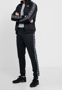 Svart träningsjacka och byxor med vita sidostricka och logotyper, tillverkade av ett slätt tyg. Modellen bär svarta sportskor.