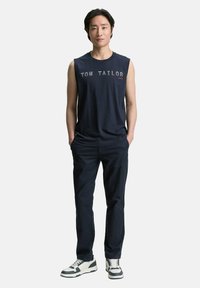 T-shirt senza maniche in cotone blu Navy con grafica "TOM TAILOR 1982", abbinata a pantaloni scuri e sneakers bianche con dettagli neri.