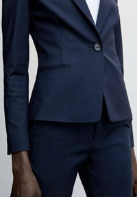 Costume bleu marine sur mesure comprenant un blazer ajusté avec une fermeture à un bouton, des revers marqués et des détails de couture contrastants ; associé à un pantalon slim.