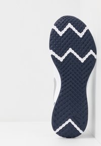 Suela de una zapatilla deportiva en blanco y negro con un patrón de relieve en zigzag, posicionada verticalmente contra una pared blanca en la esquina.