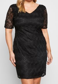 Svart spetsklänning med V-ringning, korta ärmar och texturerat zigzag-mönster. Figursydd siluett, knälång fåll. Slät foder under.