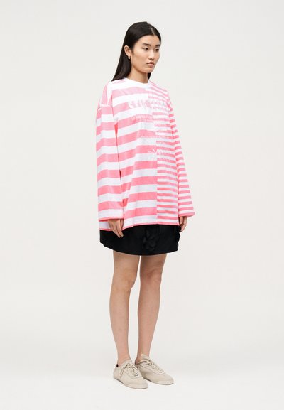 Νεαρή γυναίκα στέκεται φορώντας μια υπερμεγέθη ροζ και λευκή ριγέ μπλούζα με μακριά μανίκια, μαύρη φούστα με floral λεπτομέρειες και μπεζ αθλητικά παπούτσια.