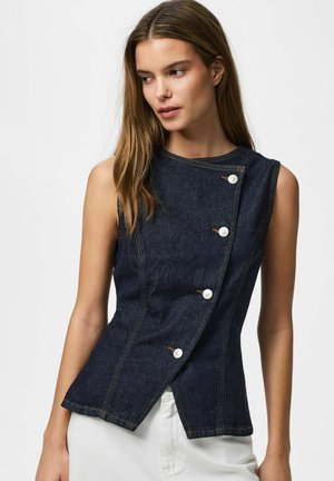 Donna che indossa un gilet di jeans scuro senza maniche con bottoni bianchi asimmetrici, abbinato a pantaloni bianchi, su sfondo semplice.