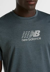 Harmaa urheilu t-paita, joka on valmistettu hengittävästä kankaasta, jossa on teksturoidut kuviot ja suuri valkoinen "New Balance" -logo edessä.