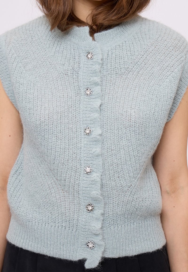 Femme portant un cardigan sans manches tricoté bleu clair avec cinq boutons argentés en forme de fleur sur le devant.