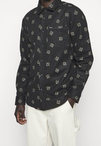 Zwart overhemd met knopen en een gestructureerde stof met een herhalend lichtgekleurd bloemmotief, een borstzak en een klassieke kraag.