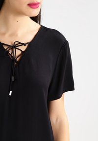 Blouse noire à manches courtes et col en V avec un laçage à l'avant. Texture douce et accents argentés minimalistes.
