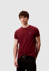 Bordeauxrotes Baumwoll-T-Shirt mit aufgerollten Ärmeln und kleinem, gesticktem Logo auf der Brust, kombiniert mit dunklen Jeans und einem schwarzen Gürtel.