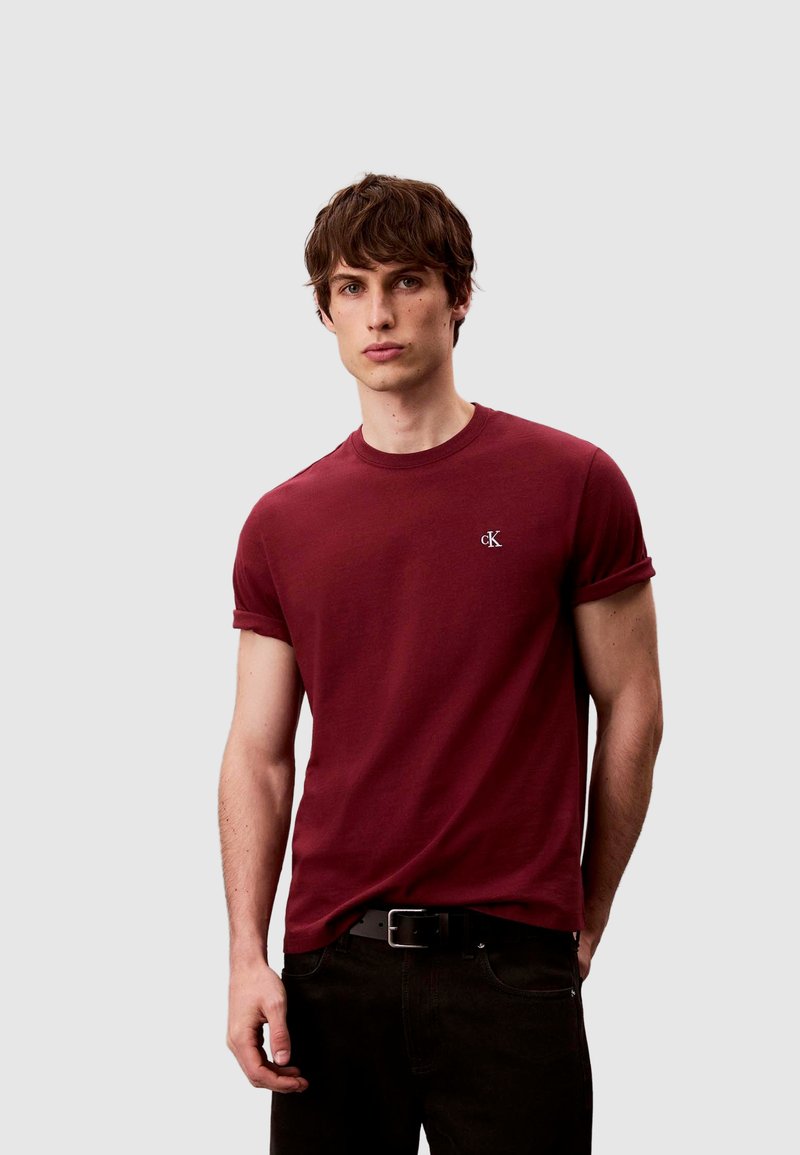 Bordeauxrotes Baumwoll-T-Shirt mit aufgerollten Ärmeln und kleinem, gesticktem Logo auf der Brust, kombiniert mit dunklen Jeans und einem schwarzen Gürtel.