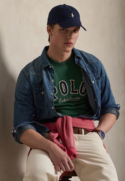 Τζιν πουκάμισο πάνω από πράσινο T-shirt με τυπώματα «Polo», ροζ φούτερ δεμένο στη μέση, παντελόνι σε εκρού απόχρωση και ναυτικό καπέλο με λογότυπο. Καφέ ρολόι και ζώνη.
