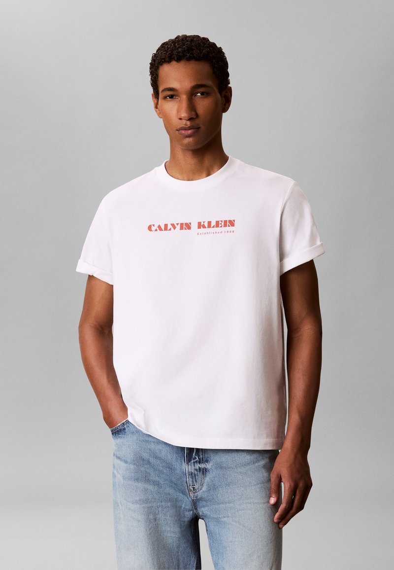 Homme portant un T-shirt blanc à manches courtes avec le texte rouge "Calvin Klein" et un jean bleu clair, debout devant un fond gris uni.